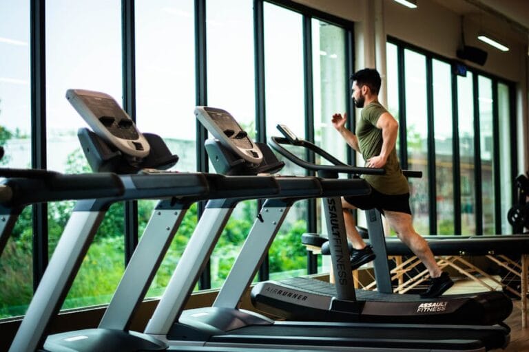 Young Man Workouts On Treadmill In Modern Gym With Large Windows And Natural Light.